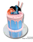 Two-tiered cake with pink and blue icing, topped with chocolate bars and flowers on a white background.