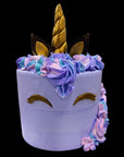 A decorated unicorn-themed cake with a light lavender body, gold horn and closed-eye details, and purple, blue, and pink piped frosting accents on a black background.