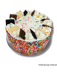 A round vanilla sponge celebration cake with white frosting, colorful sprinkles, piped cream decorations, and chocolate and white chocolate bar pieces on top, shown on a silver cake board against a white background.
