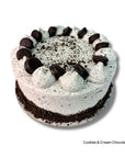 A round chocolate cake with white cream frosting, topped with piped dollops of frosting, chocolate cookie crumbs, and chocolate cookie pieces, on a white background with text reading 'Cookies & Cream Chocolate Cake' in the bottom right corner.