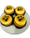 Four cupcakes with yellow icing and 'RU OK?' text on a white plate.