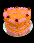 Two-tiered cake with pink and orange frosting, topped with 'Gemini Baby 34' Love heart shaped