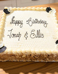 A square birthday cake with white piped frosting, almond shavings around the base, and black decorative corner accents. The top is written with black icing reading 'Happy Birthday Jonah & Ellis' on a wooden table.