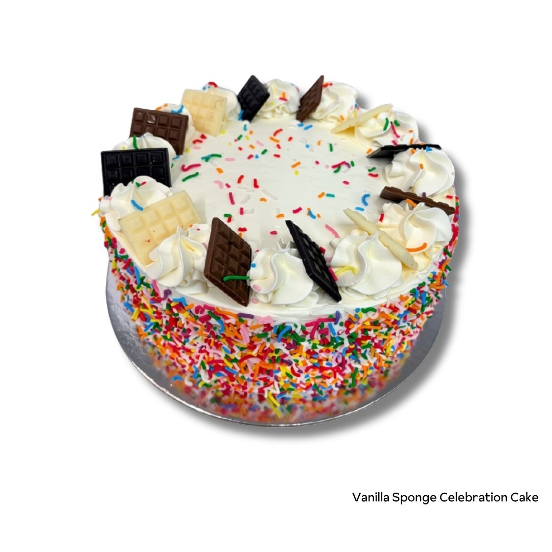 A round vanilla sponge celebration cake with white frosting, colorful sprinkles, piped cream decorations, and chocolate and white chocolate bar pieces on top, shown on a silver cake board against a white background.