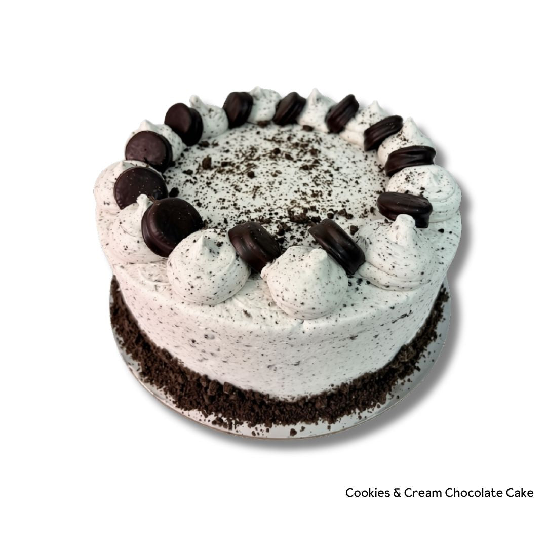 A round chocolate cake with white cream frosting, topped with piped dollops of frosting, chocolate cookie crumbs, and chocolate cookie pieces, on a white background with text reading 'Cookies &amp; Cream Chocolate Cake' in the bottom right corner.
