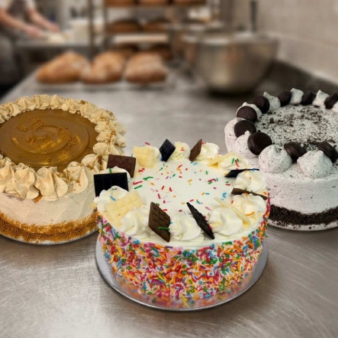 Three decorated cakes displayed on a stainless steel counter in a bakery setting. The center cake is round with white whipped cream, colorful sprinkles, and chocolate pieces.
