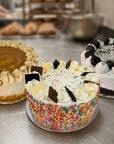 Three decorated cakes displayed on a stainless steel counter in a bakery setting. The center cake is round with white whipped cream, colorful sprinkles, and chocolate pieces.
