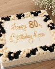 A large square birthday cake with cream-colored frosting, decorated with black and cream rosette borders and gold lettering that reads 'Happy 80th Birthday Ross', shown on a wooden surface with small white flowers in the corner.