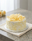 A round red velvet cake with white chocolate decorations, decorated with a large rosette on top and white chocolate shavings on the sides, sitting on a beige napkin on a speckled countertop.